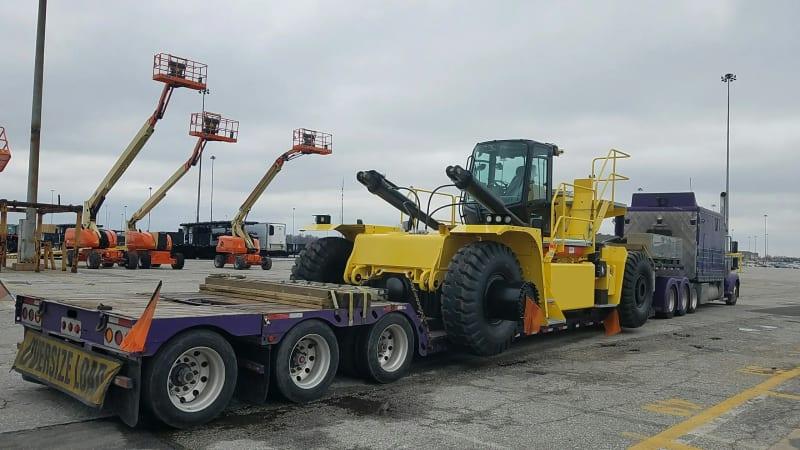 A truck carries a yellow forklift through a parking lot.