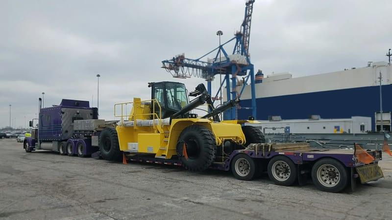 The rear of a purple truck carrying a yellow forklift through a parking lot.