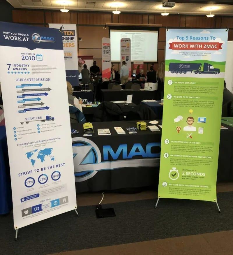 A booth for ZMac at a college career fair, with a tablecloth and two banners detailing job benefits.