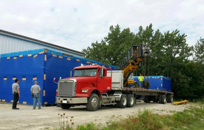A red truck parked in front of blue shipping containers with construction equipment in the truck bed.