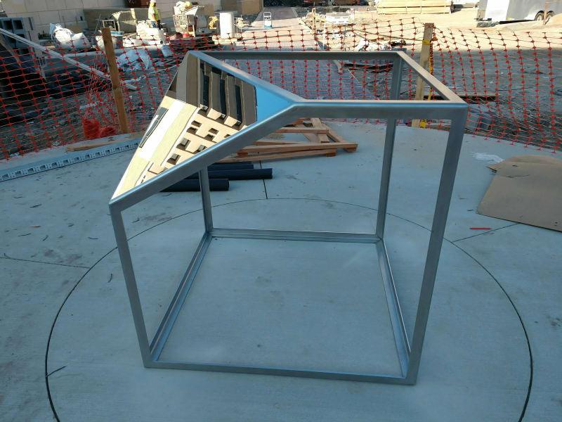 A cube-like structure of metal and mirrors sits at a construction site.