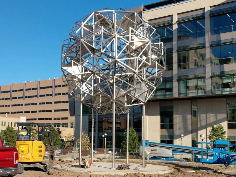 A completed sculpture of mirrors and metal forming a sphere out of various cube shapes. The sculpture sits outside a commercial building.