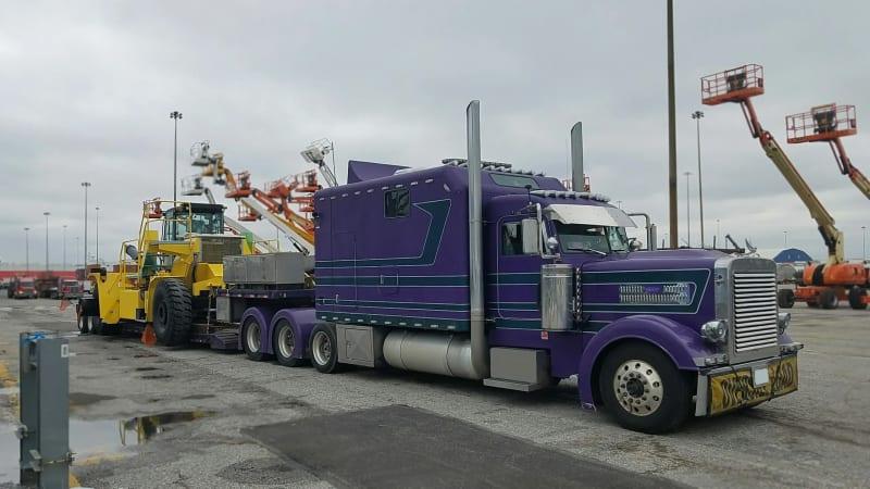 The front of a purple truck carrying a forklift in a parking lot.