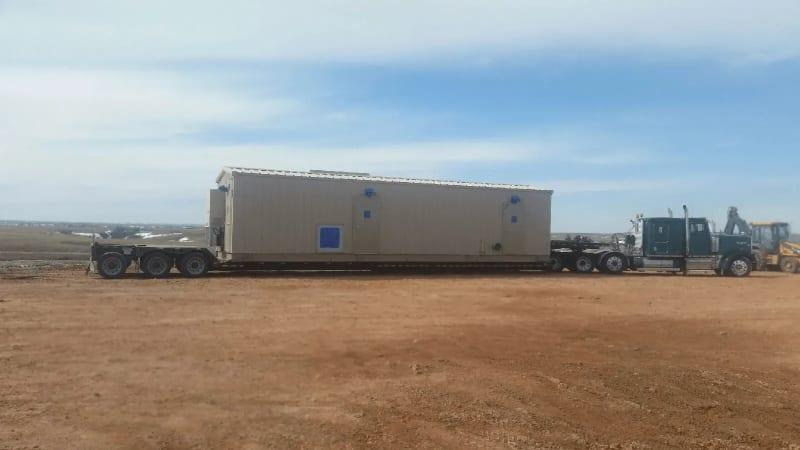 A tan prefabricated building sitting on a truck bed in a dirt lot.