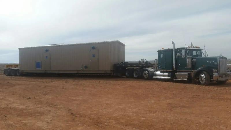 The side of the truck carrying a tan prefabricated building in a dirt lot.
