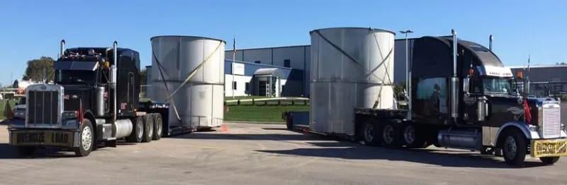 Two white tanks stand upright on the back of two overdimensional trucks.