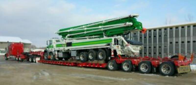 A green truck mounted concrete pump loaded onto a larger red truck.