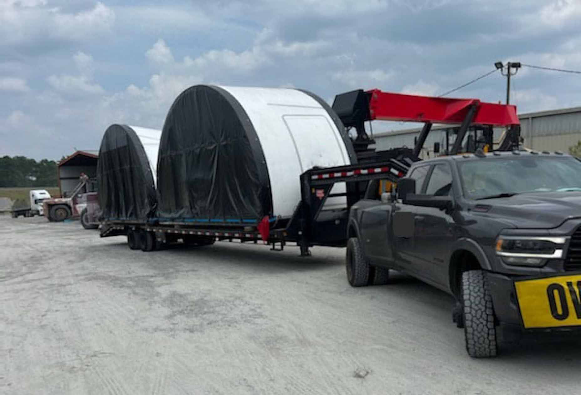 A low bed trailer attached to a pickup truck carrying two large metal half-circle containers covered in black tarp.