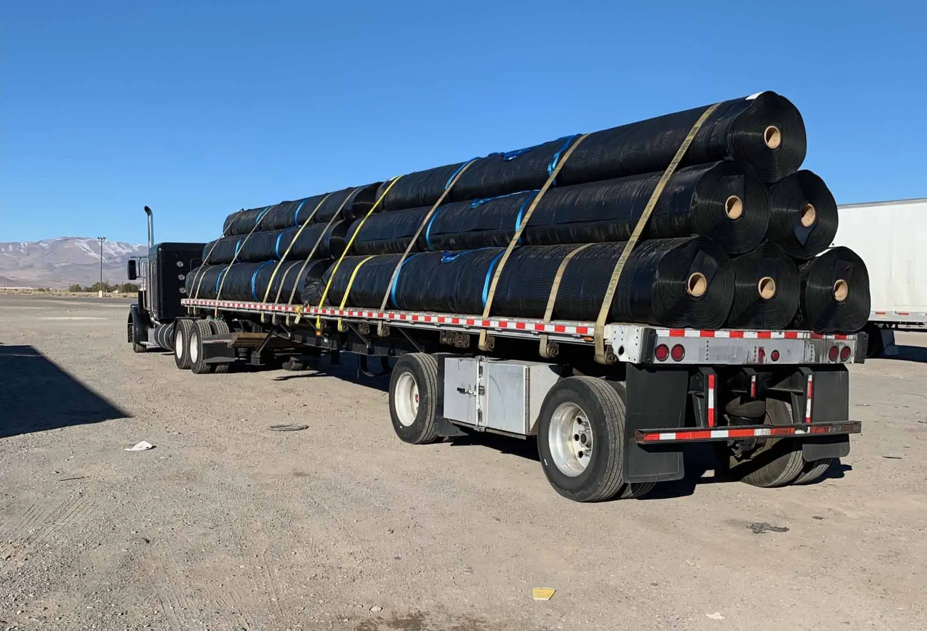 The rear of a truck with several long rolls of black tarp strapped to the bed. It is parked in a lot with a blue sky background.