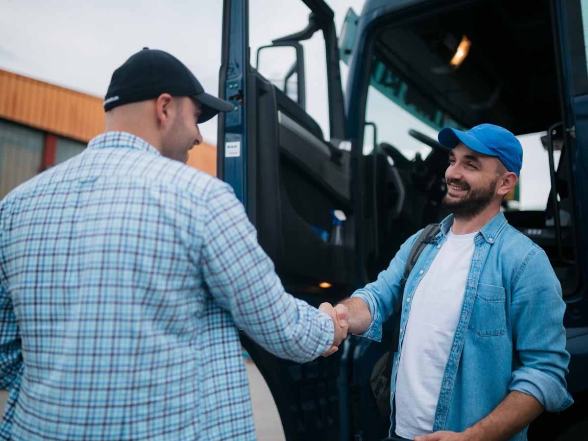 Two men wearing hats and casual attire smile and shake hands in front of a truck's open door.