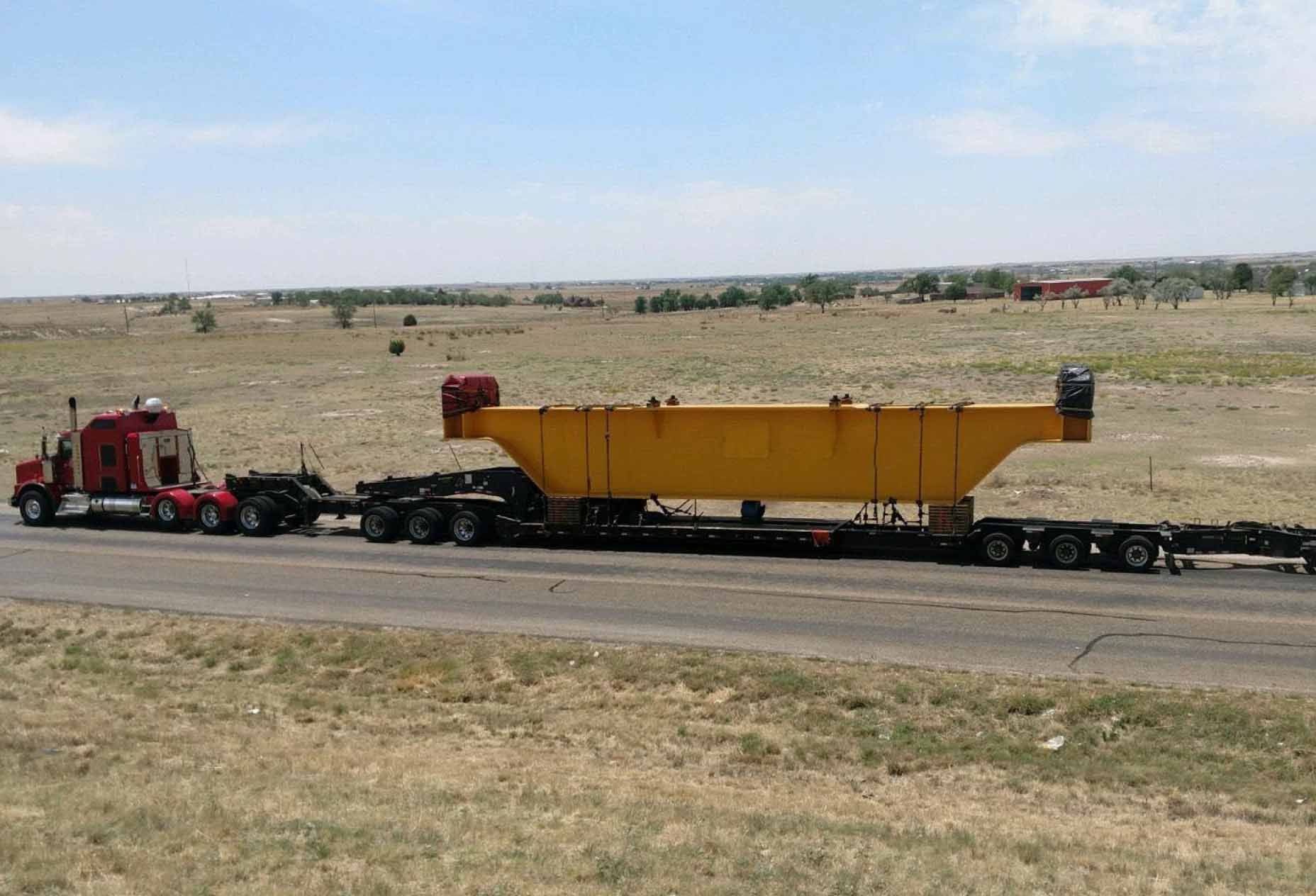 An oversized freight truck with a low bed carrying a large yellow metal piece travels down a highway surrounded by fields.