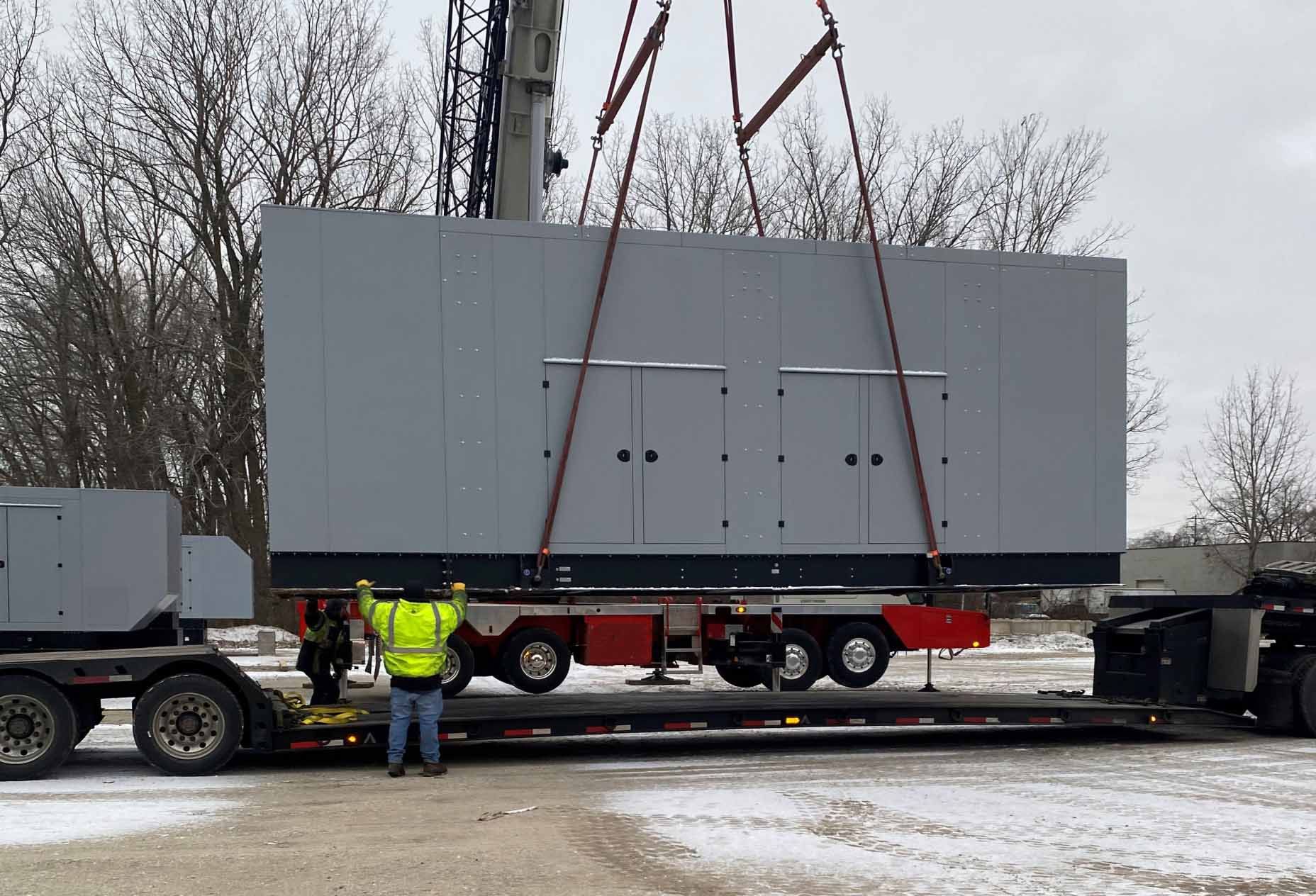 Two ZMac carriers assist the lift of a large metal box from a low truck bed using a crane.