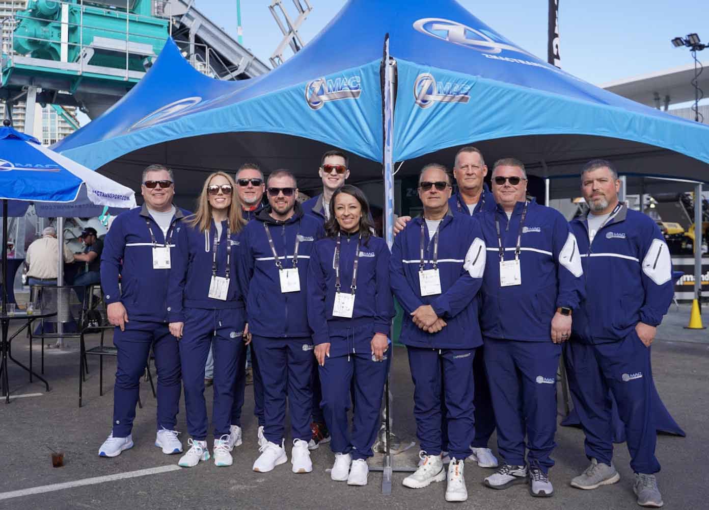 Members of the ZMac team wearing matching ZMac zip-up windbreakers stand smiling in front of an outdoor ZMac booth tent.