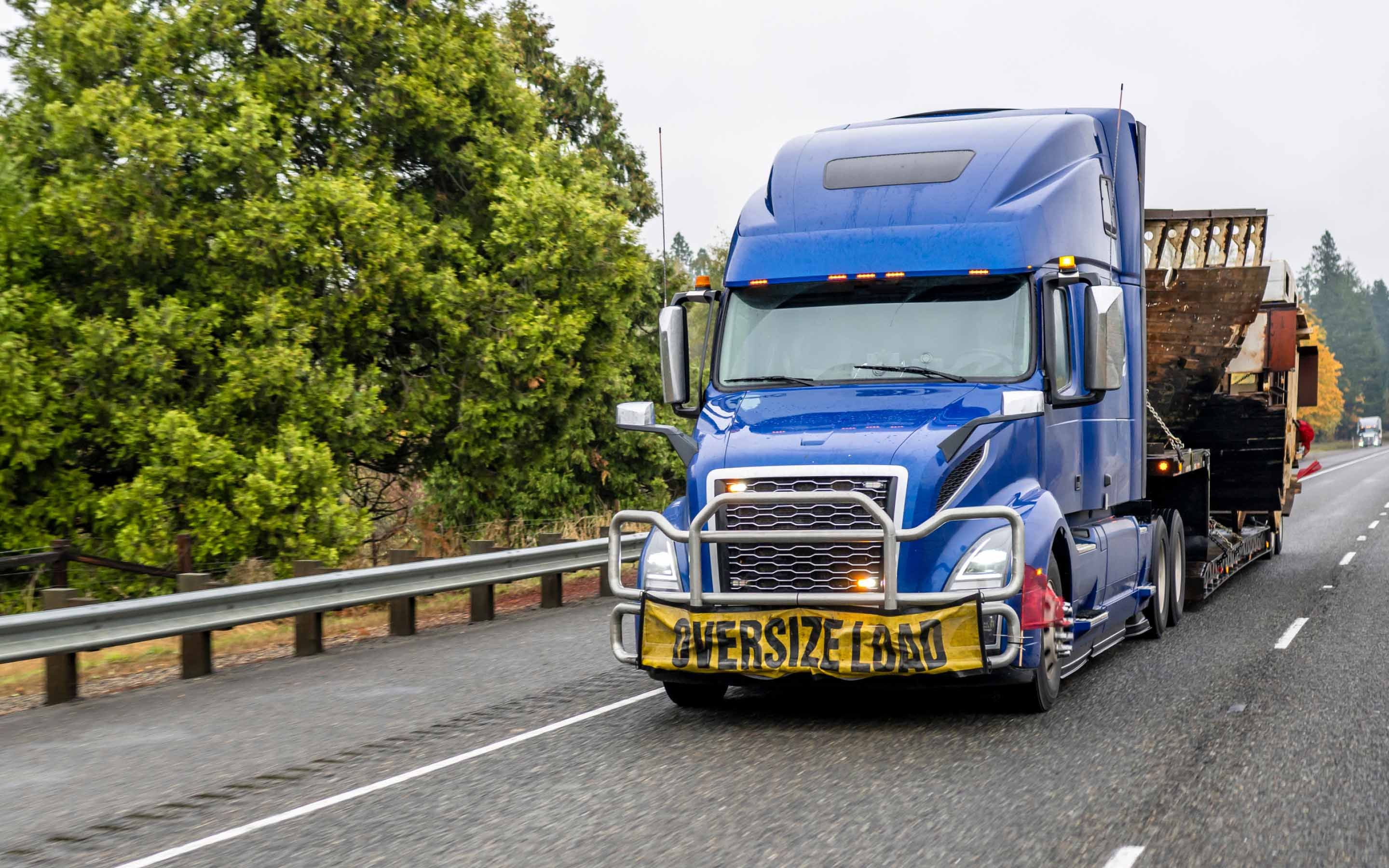 A blue overdimensional freight truck drives down a road with trees in the background.